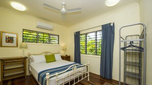 A bedroom with a bed, bedside tables, a ceiling fan, windows with curtains, and a metal drying rack. - Manyana (Nelly Bay)