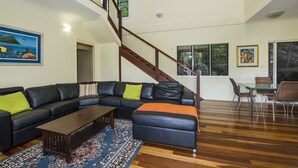 A modern living room with a black leather sectional sofa, a wooden coffee table, and a glass dining table with chairs. A staircase with wooden railings leads to an upper level. - Manyana (Nelly Bay)