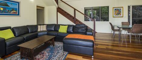 A modern living room with a black leather sectional sofa, a wooden coffee table, and a glass dining table with chairs. A staircase with wooden railings leads to an upper level.
