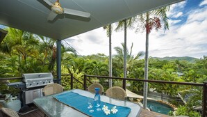 A covered outdoor dining area with a table set for two, a grill, and a view of a pool and tropical landscape. - Manyana (Nelly Bay)