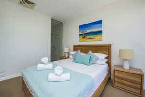 A hotel room with a bed, a painting of a beach scene, a shower, and a nightstand with a lamp.