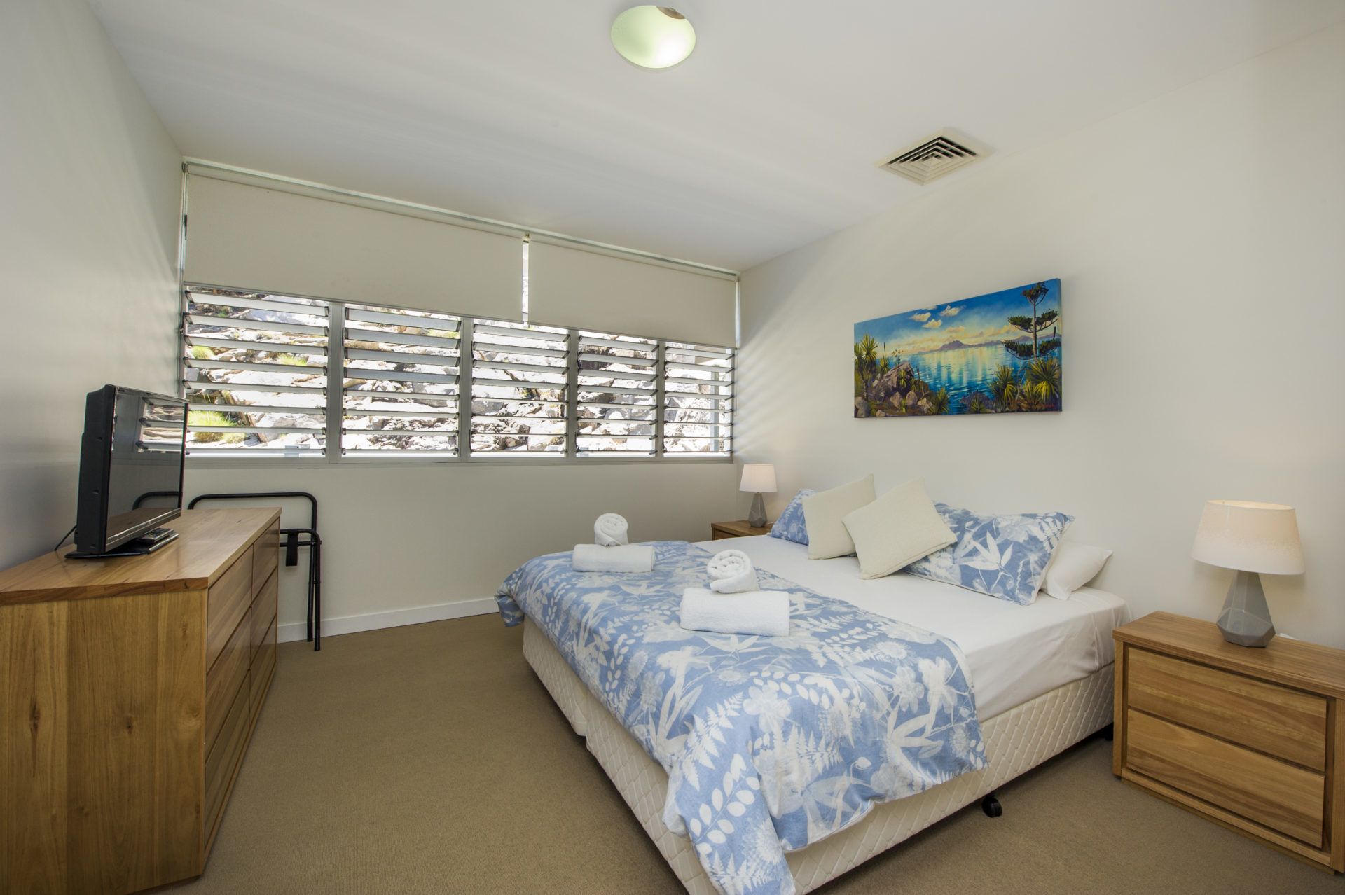 A bedroom with a bed, a desk, a chair, a window with blinds, a painting, and a lamp.