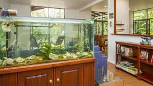 Une pièce dotée d’un grand aquarium, d’un ventilateur de plafond, d’une bibliothèque remplie de livres et offrant une vue sur la verdure par les fenêtres.