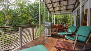 Une terrasse extérieure couverte avec un plancher en bois, des fauteuils de jardin confortables avec des coussins verts, un barbecue et une table.