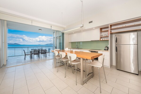 A modern kitchen with a large island, stainless steel appliances, and a view of the sea through a glass door.