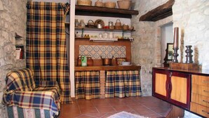 A rustic kitchen with a plaid-covered sofa, wooden shelves with kitchenware, and a stone wall. - Villa Nicetta (San Fratello)