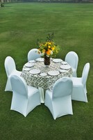 A round table with a patterned tablecloth, surrounded by white chairs, set on a grassy lawn.