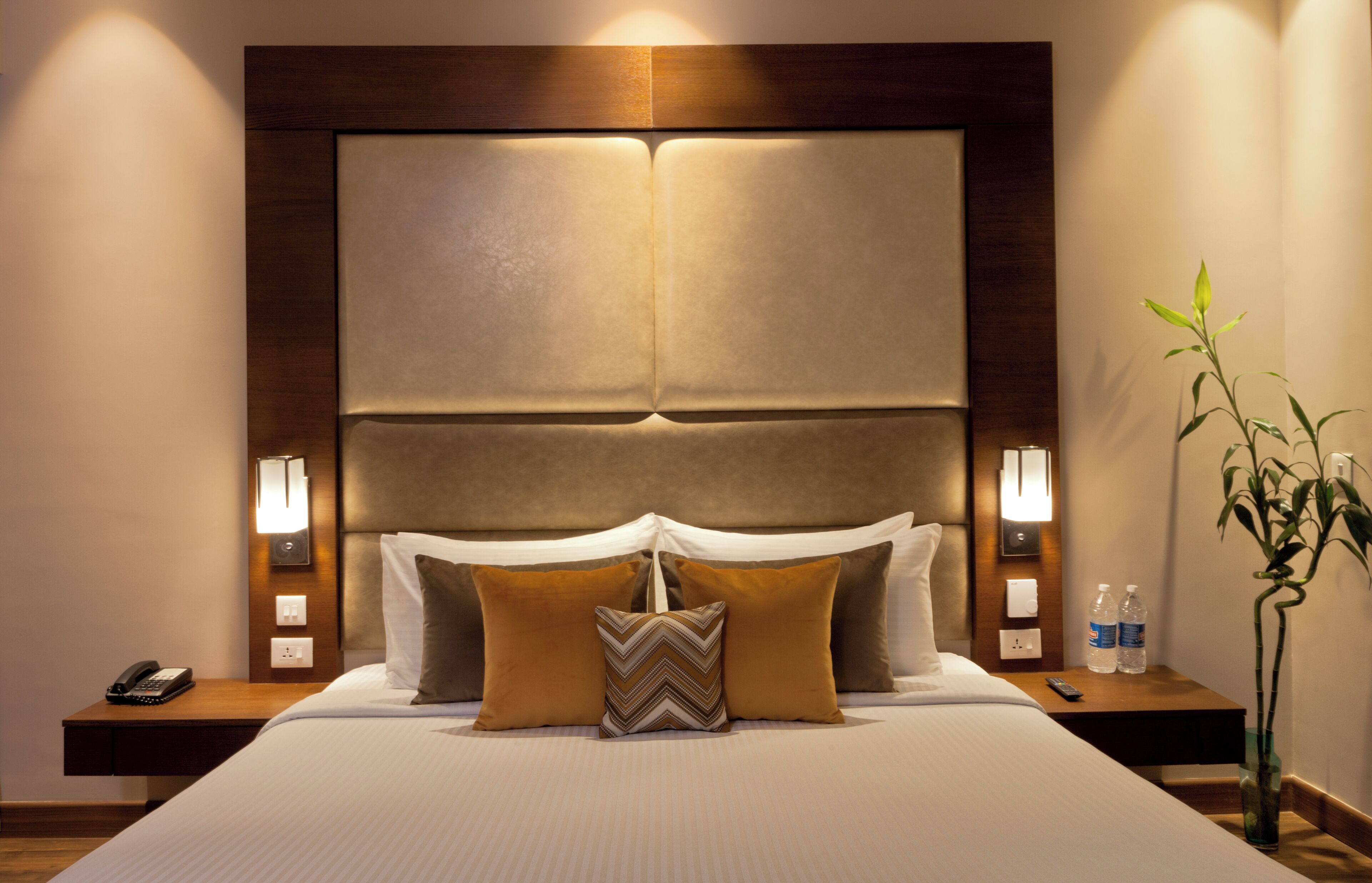 A hotel room with a large bed, two bedside tables, a nightstand, a wall-mounted lamp, and a potted plant.