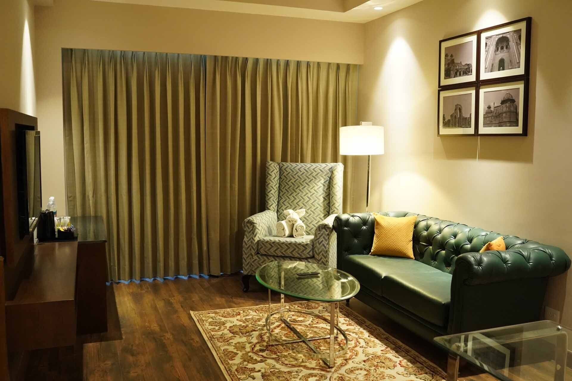 Un salon avec un canapé vert, un fauteuil à motifs, une table basse en verre, une lampe et une grande fenêtre avec des rideaux.