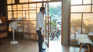 A man standing in a room with a large window, a fan, and a motorcycle outside.