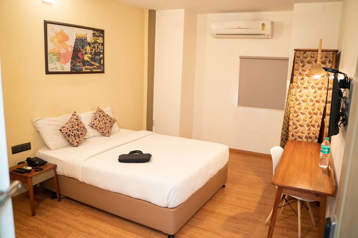 Habitación de hotel con cama, escritorio, silla, lámpara, botella de agua y un cuadro en la pared.
