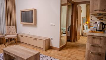 A modern living room with a sofa, a coffee table, a TV mounted on the wall, wooden flooring, and a kitchen area visible through an open door.