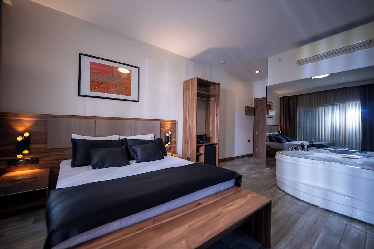 A modern hotel room with a large bed, a wooden bench, a built-in TV, a large window, and a hot tub.
