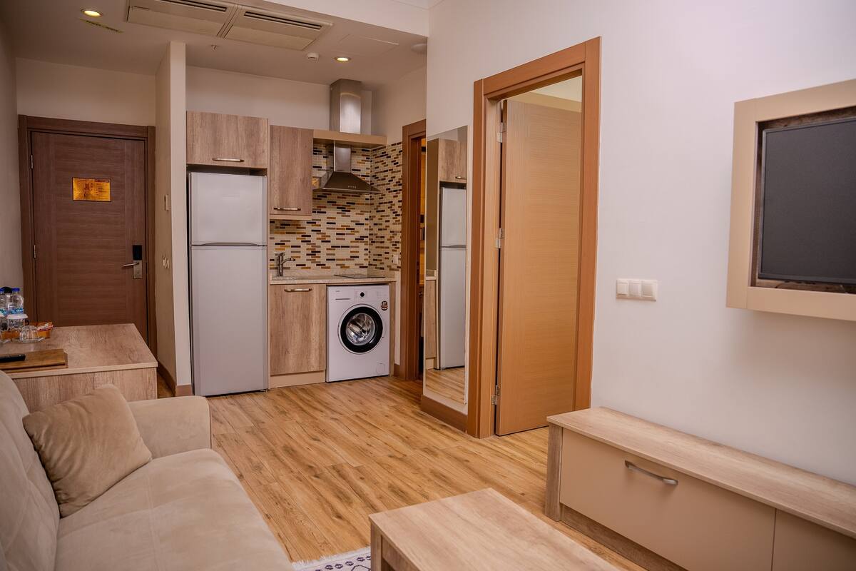 A compact living space with a kitchenette, a washing machine, a sofa, and a TV mounted on the wall.