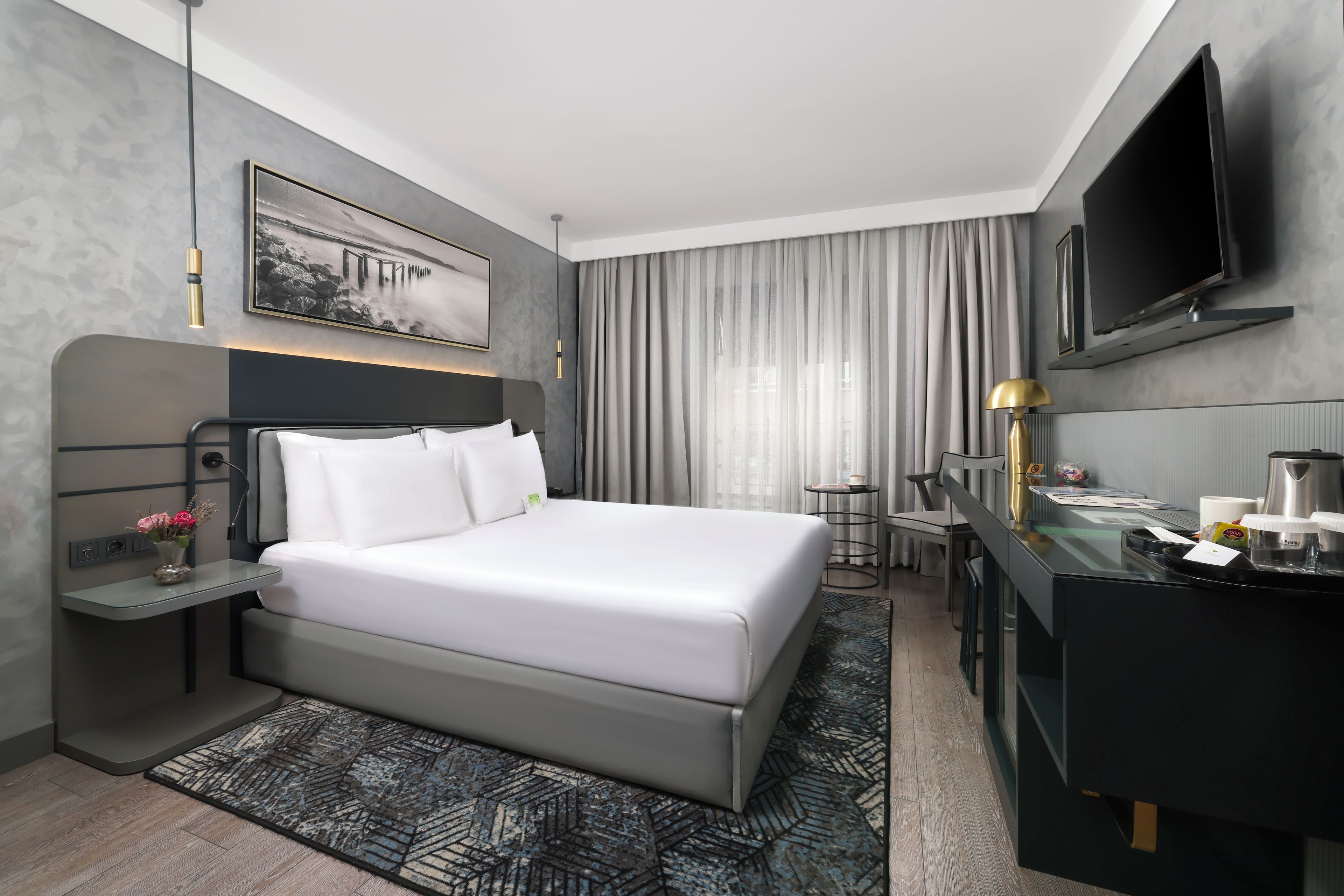 Ein modernes Hotelzimmer mit Bett, Nachttisch, Lampe, Fernseher und Schreibtisch mit Kaffeemaschine.