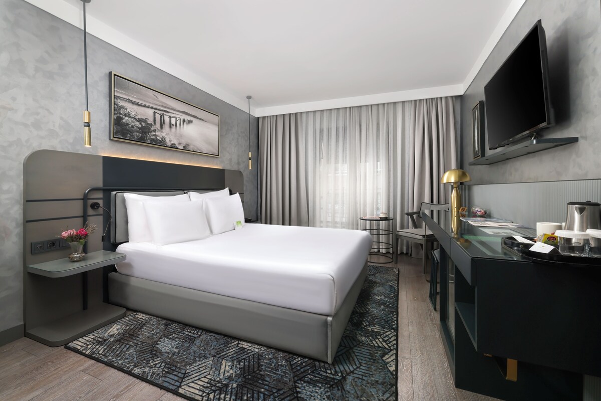 A modern hotel room with a bed, bedside table, lamp, TV, and a desk with a coffee maker.