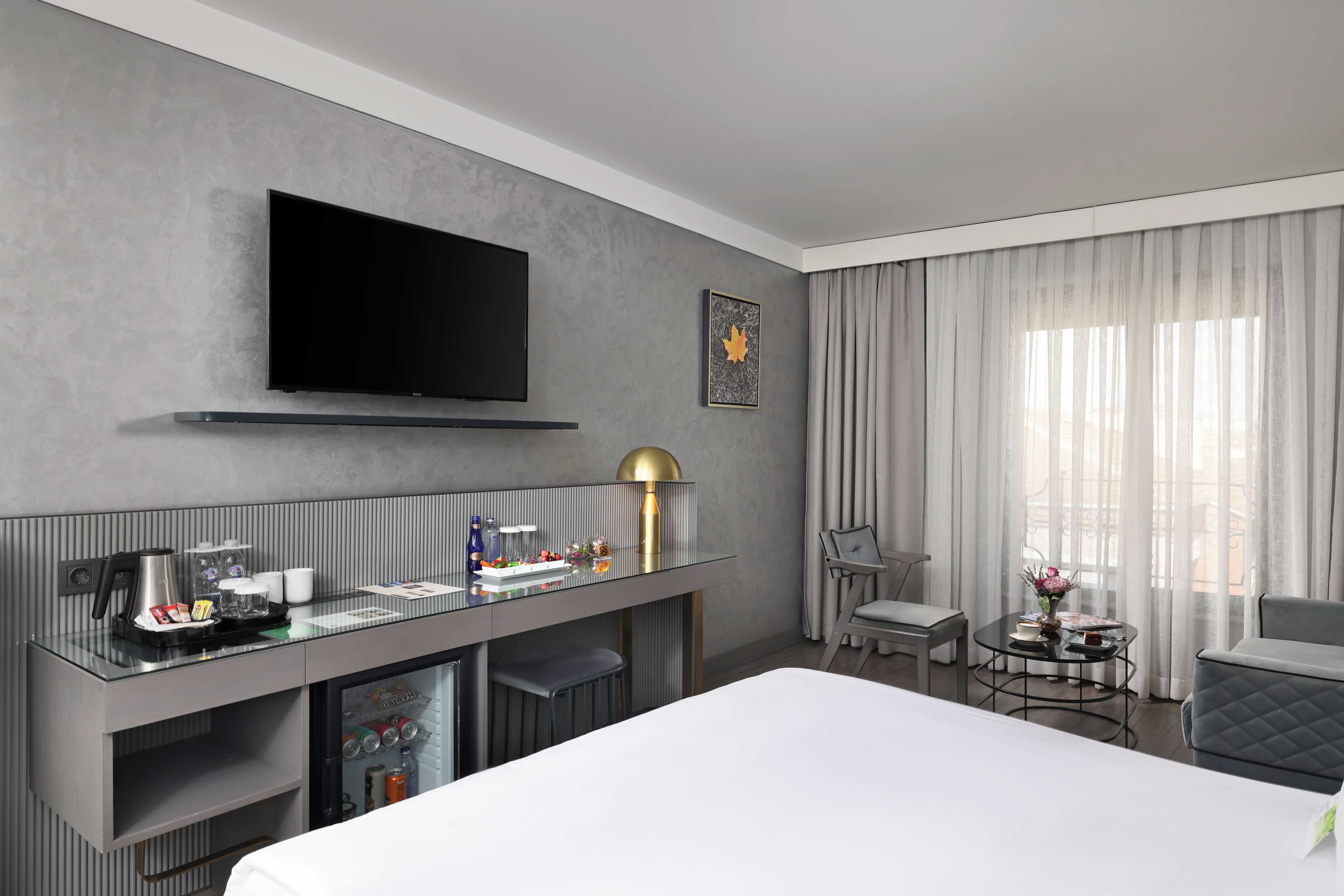 Una habitación de hotel moderna con televisor de pantalla plana, minibar, zona de estar con mesa y sillas, y un amplio ventanal con cortinas.