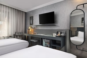 Una habitación de hotel moderna con dos camas, un escritorio, un televisor de pantalla plana, un minibar y un espejo grande.