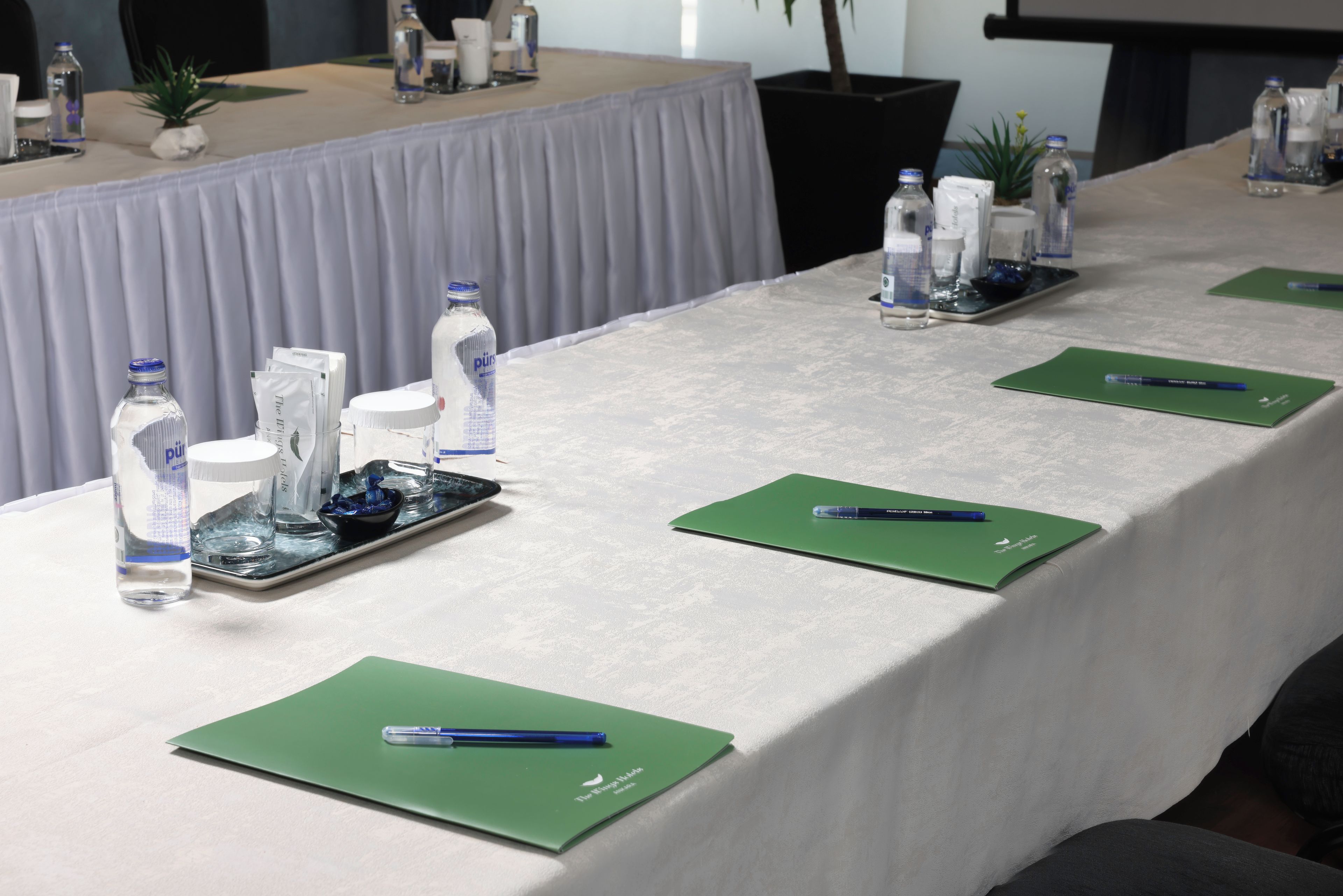 Una mesa de conferencias con botellas de agua, un bol de aperitivos y blocs de notas verdes con bolígrafos.