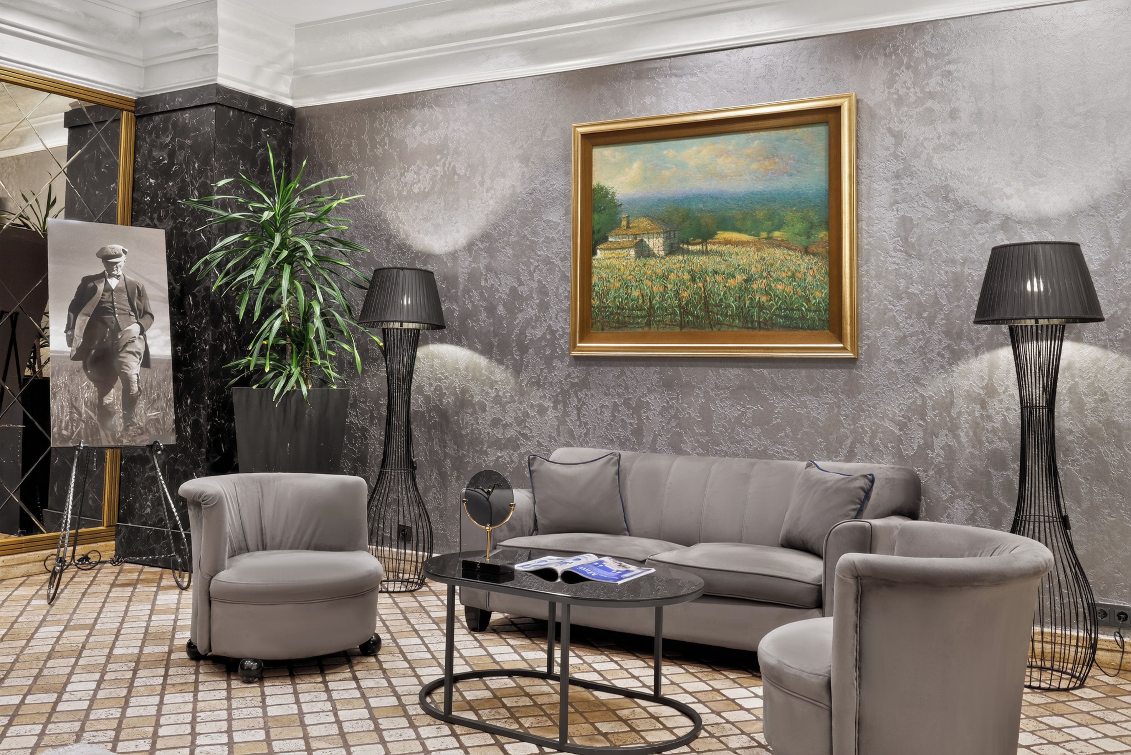 A modern living room with a grey sofa, two armchairs, a coffee table, a floor lamp, a painting, a potted plant, and a framed photograph.