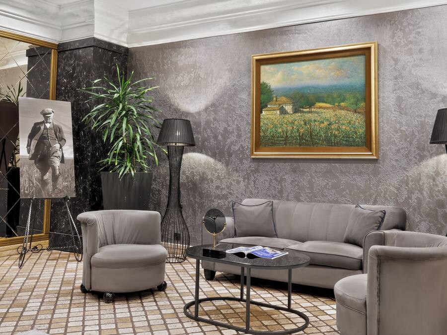 Een moderne woonkamer met een grijze bank, twee fauteuils, een salontafel, een vloerlamp, een schilderij, een potplant en een ingelijste foto.