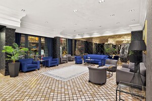 Um hall amplo com piso de azulejos, várias áreas de estar incluindo sofás e poltronas, e uma grande janela com vista para o exterior.