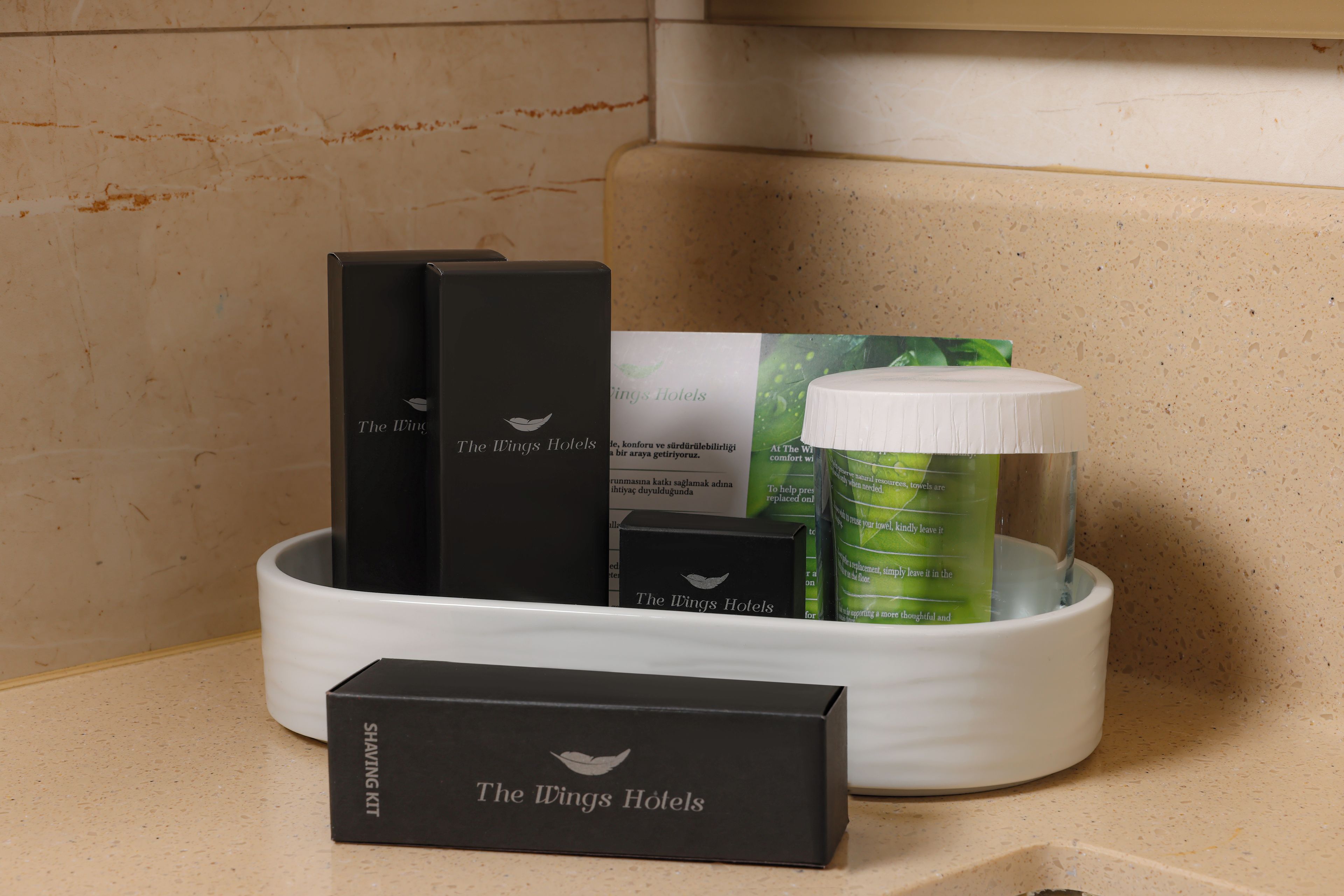 A hotel bathroom with a sink, a mirror, and toiletries including a shaving kit, a green bottle, and a white container.