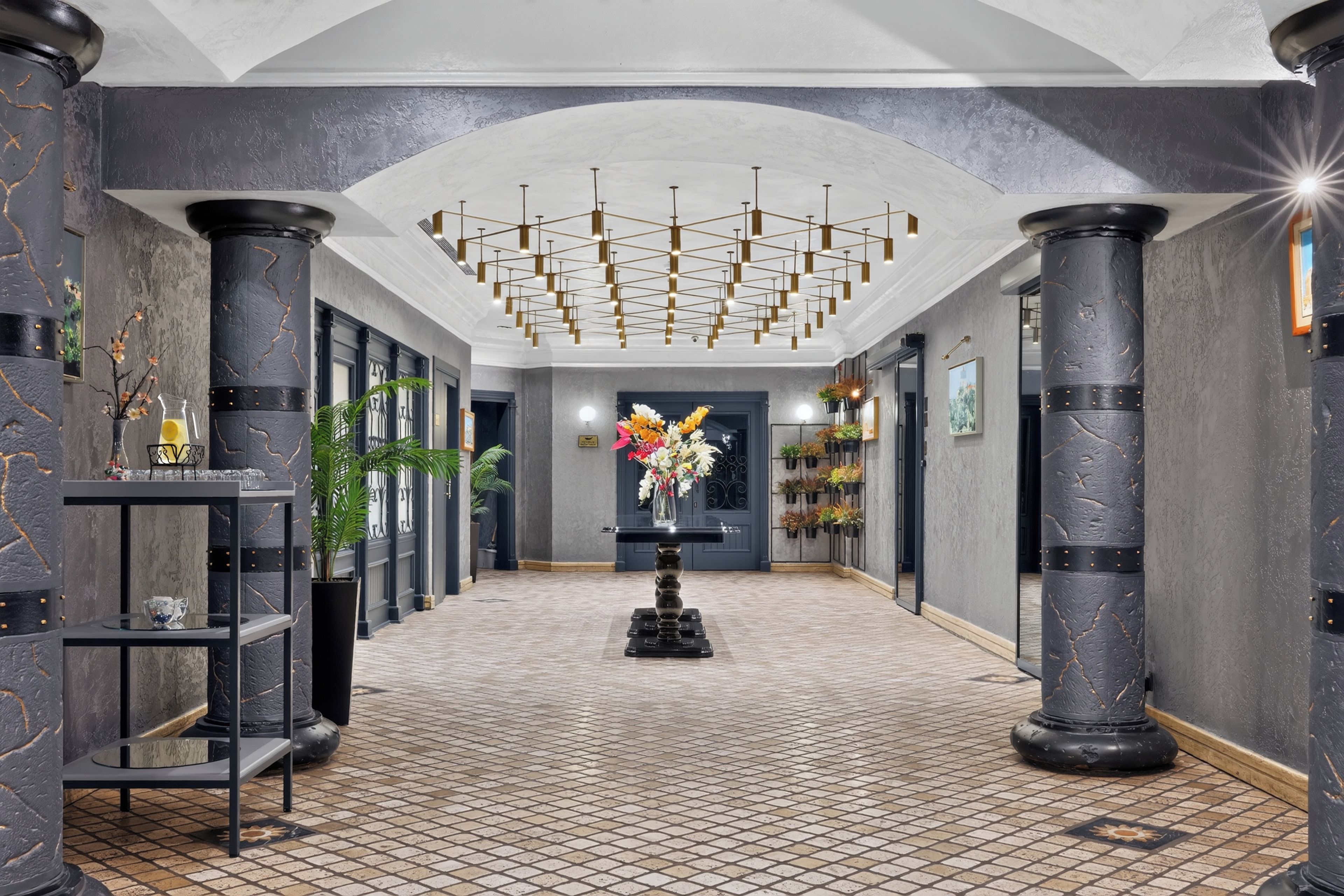 A hallway with a tiled floor, black columns, a central floral arrangement, and a ceiling with a geometric light fixture.