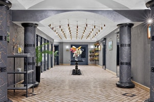 Un pasillo con piso de baldosas, columnas negras, un arreglo floral central y un techo con lámpara de diseño geométrico.