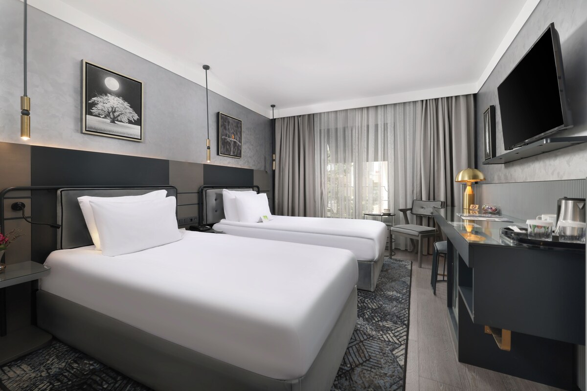 A hotel room with two beds, a desk, a TV, and a window with curtains.