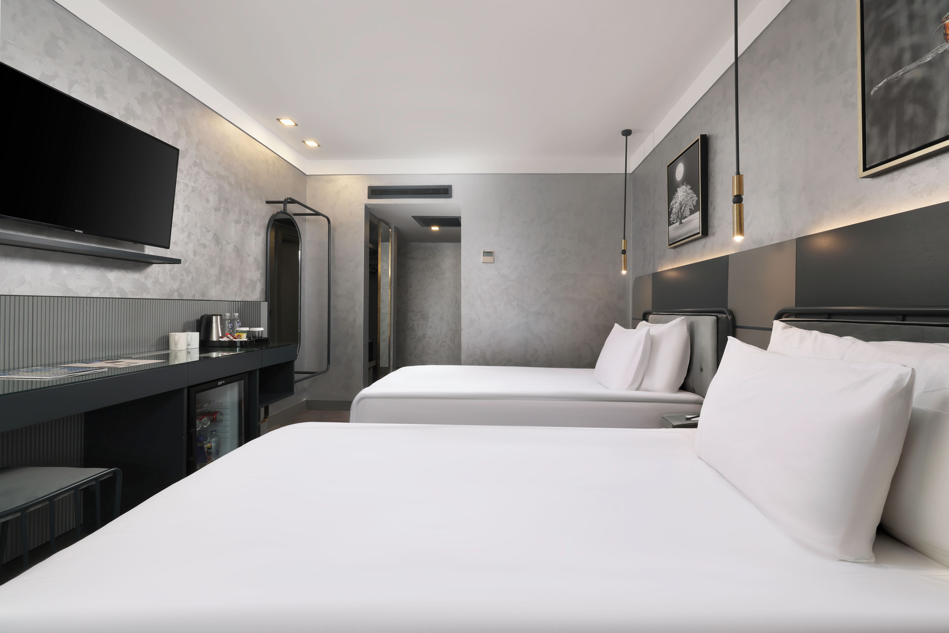A modern hotel room with two beds, a flat-screen TV, a minibar, and a large mirror.
