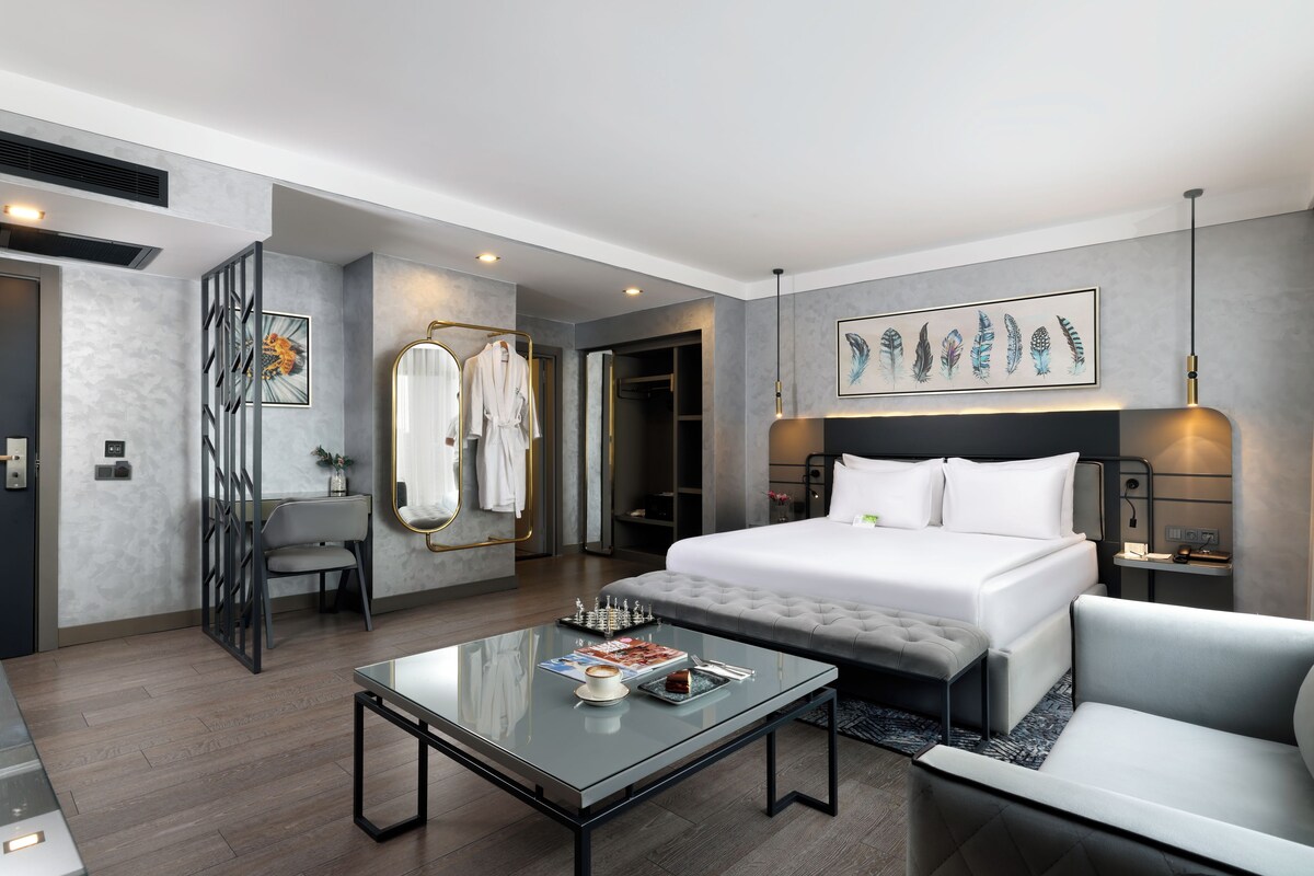A modern hotel room with a large bed, a glass coffee table, a sofa, and a painting on the wall.