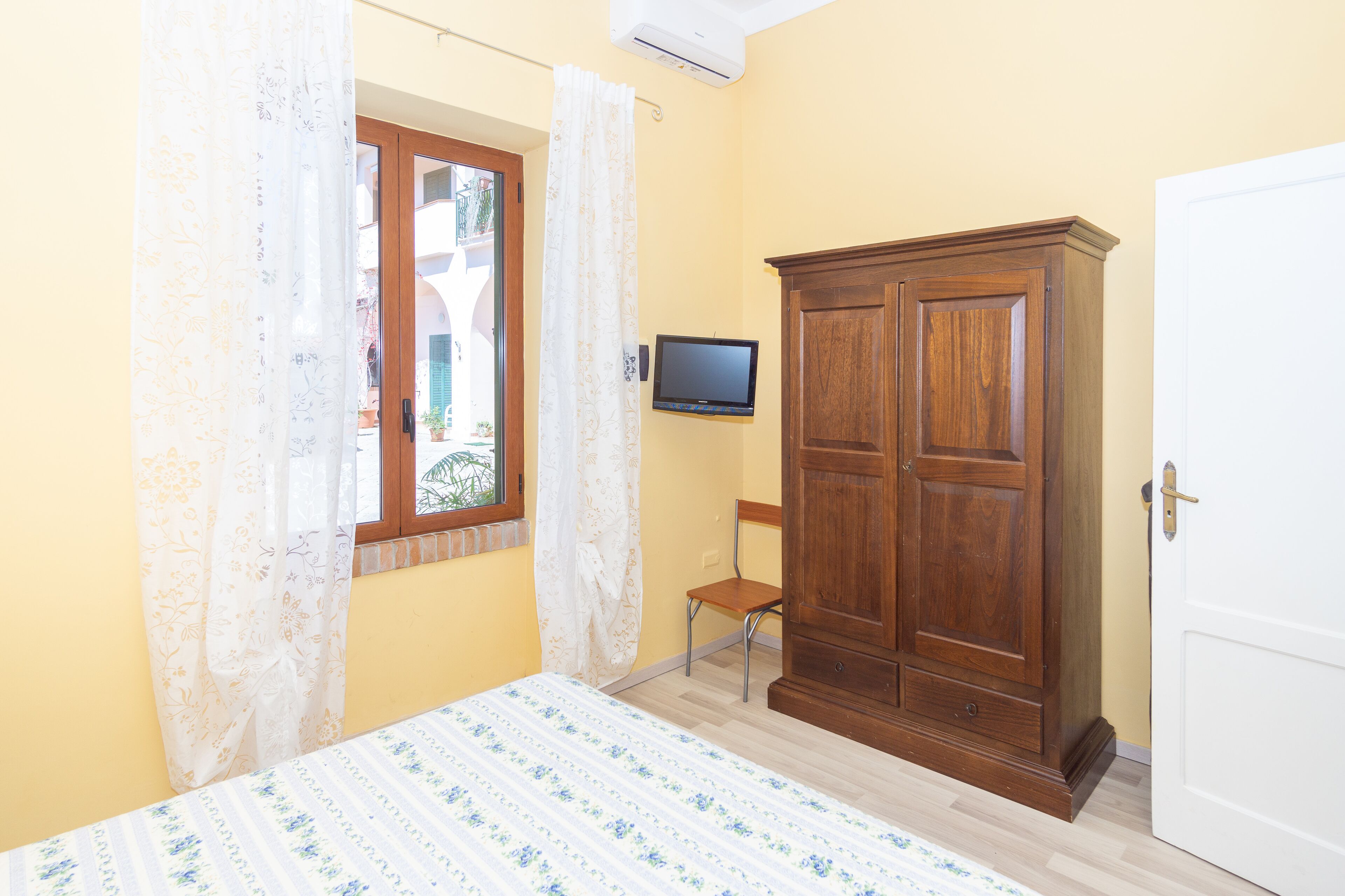 A bedroom with a bed, a wooden wardrobe, a television, and a window with curtains.