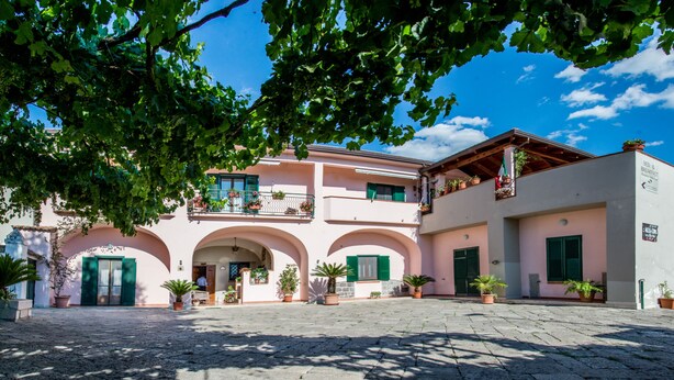 A building with a pink facade, green shutters, and multiple balconies. There are potted plants and a sign indicating a breakfast area. - La Masseria (Capua)