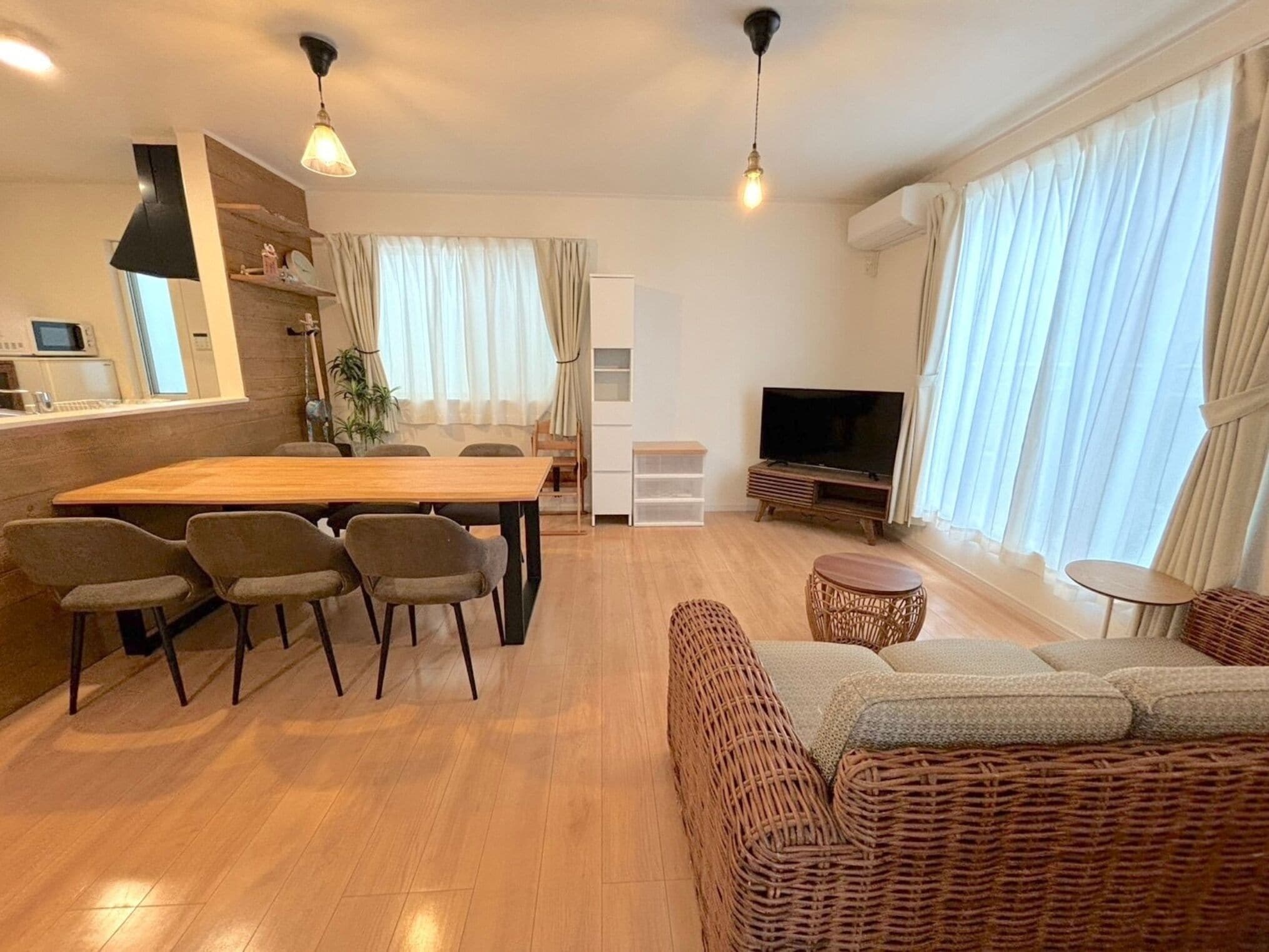 A modern living room with a wooden dining table, four chairs, a sofa, a TV, and a kitchen area with a microwave.