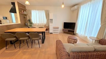 A modern living room with a wooden dining table, four chairs, a sofa, a TV, and a kitchen area with a microwave.