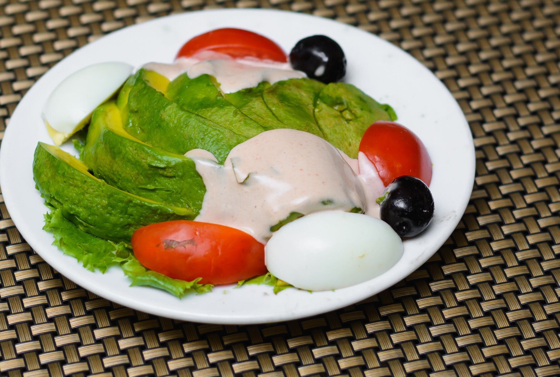 A plate of salad with lettuce, tomatoes, olives, and a white egg on a woven tablecloth.