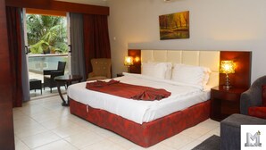 A hotel room with a large bed, two bedside tables with lamps, a chair, a balcony with a table and chairs, and an air conditioning unit. - Mendiata Hotel (Accra)