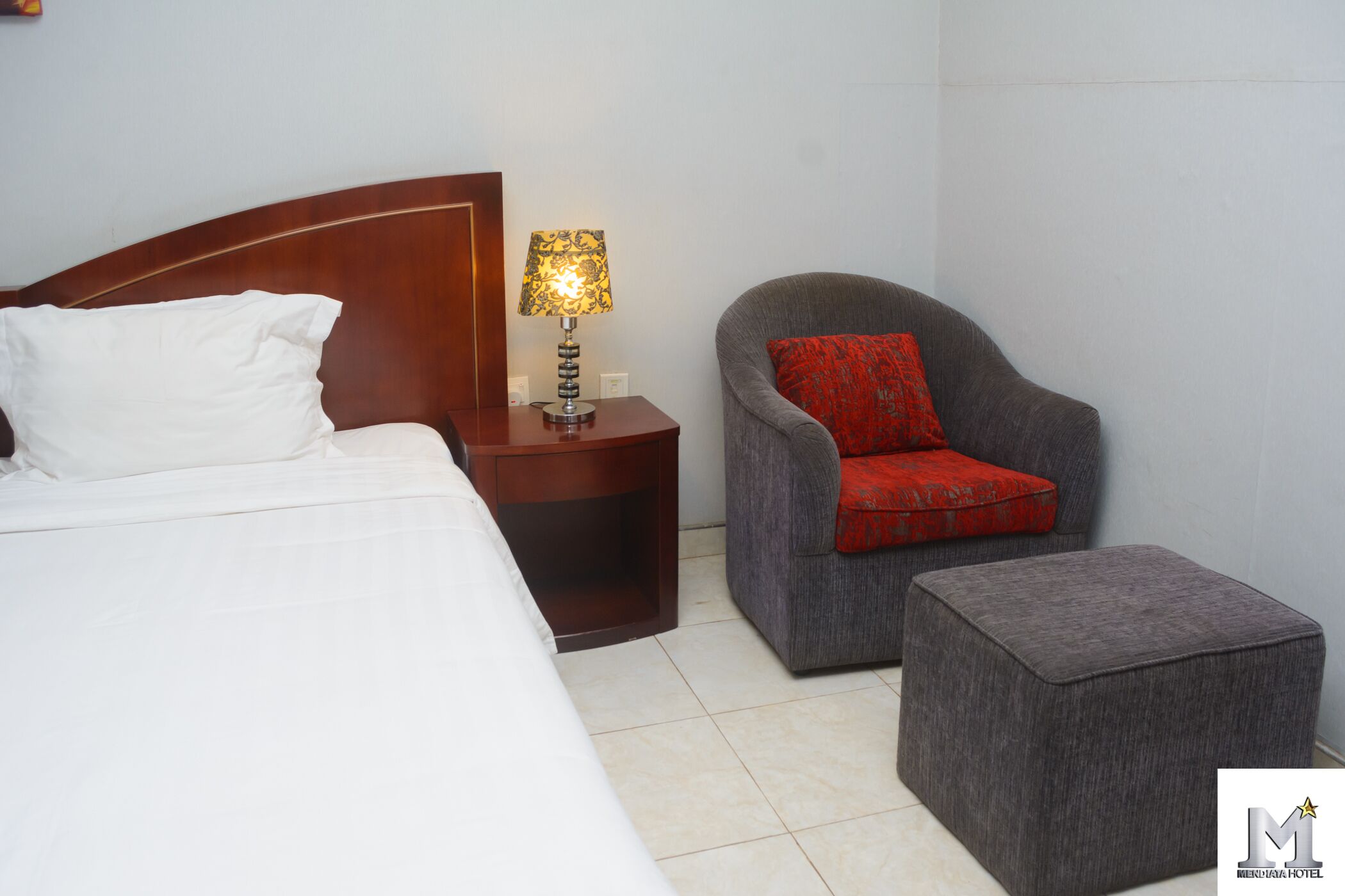 Una habitación de hotel con una cama, una mesita de noche con lámpara, un sillón gris con cojín rojo y un otoman gris.