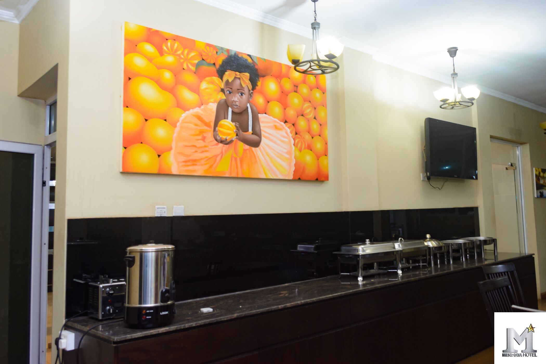 A kitchen with a large painting of oranges, a coffee maker, and a flat-screen TV on the wall.