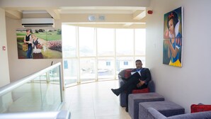 A waiting area with a glass railing, a man sitting on a bench, and two paintings on the wall. - Mendiata Hotel (Accra)
