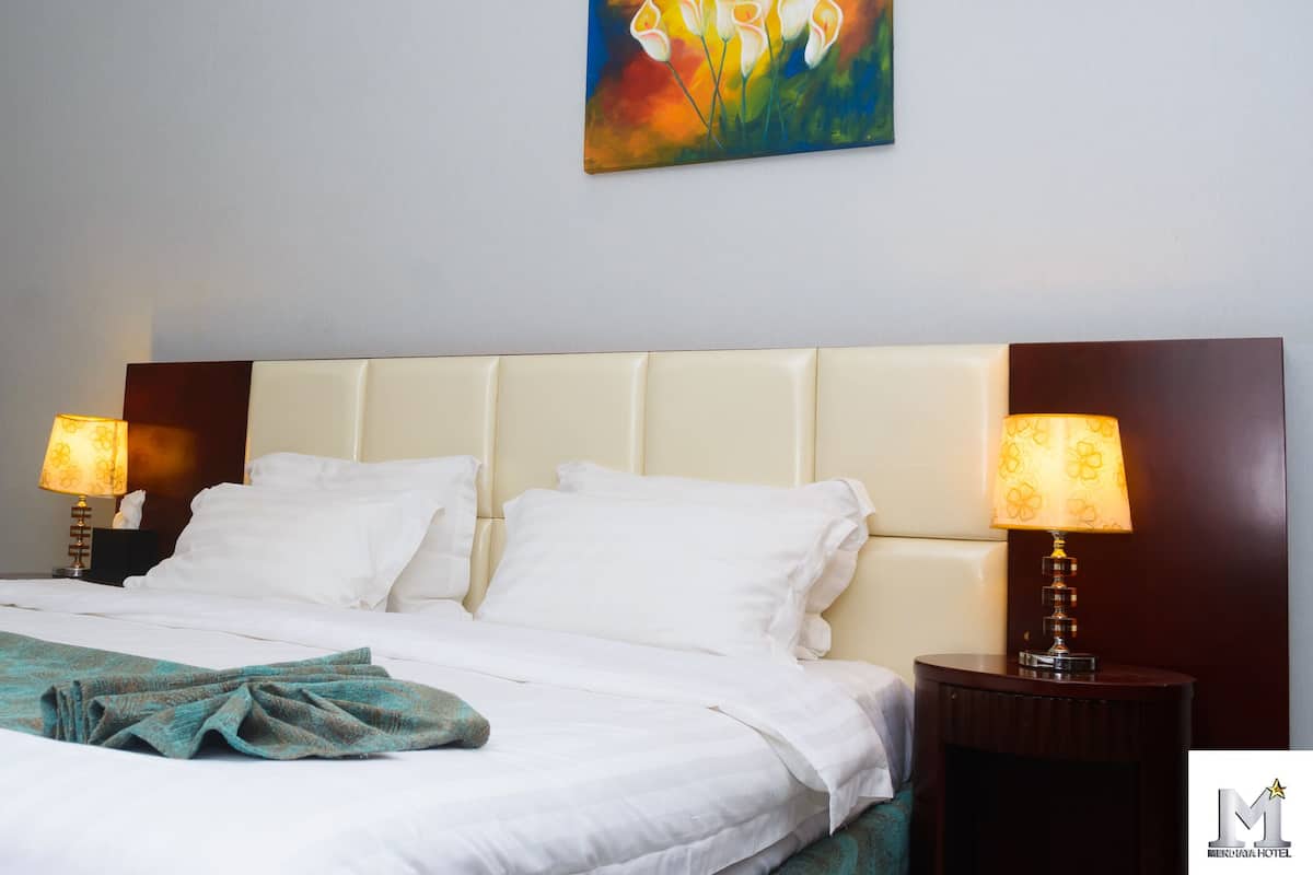 A hotel room with a bed, two bedside lamps, a painting on the wall, and a logo in the corner.