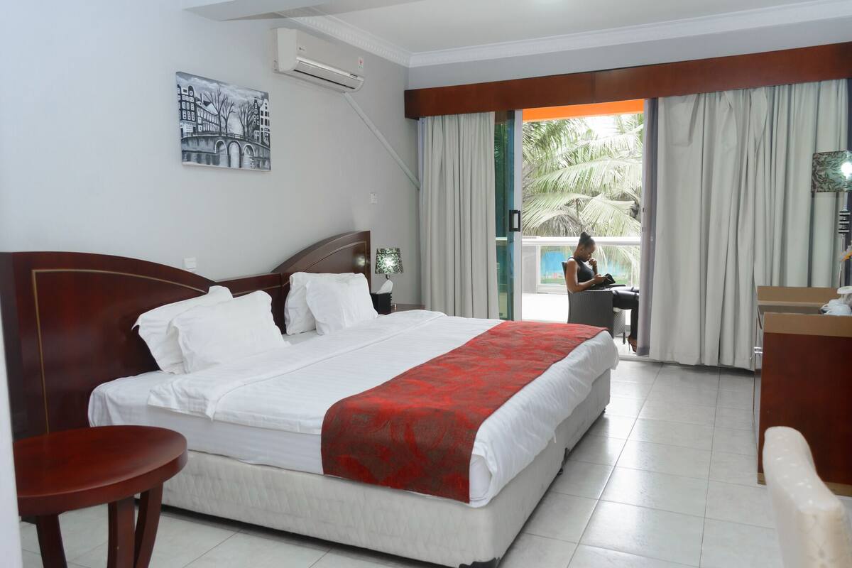 A hotel room with a large bed, a desk, a chair, a window with curtains, and a painting on the wall.