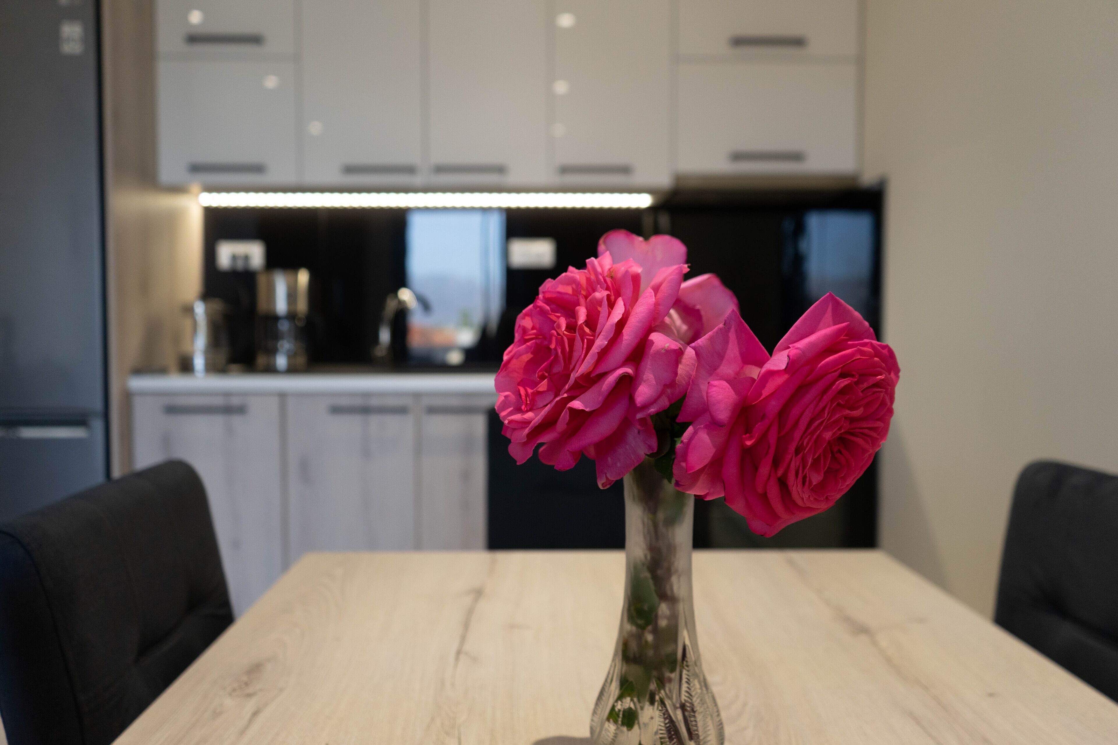 A dining table with a vase of pink flowers and a modern kitchen in the background.