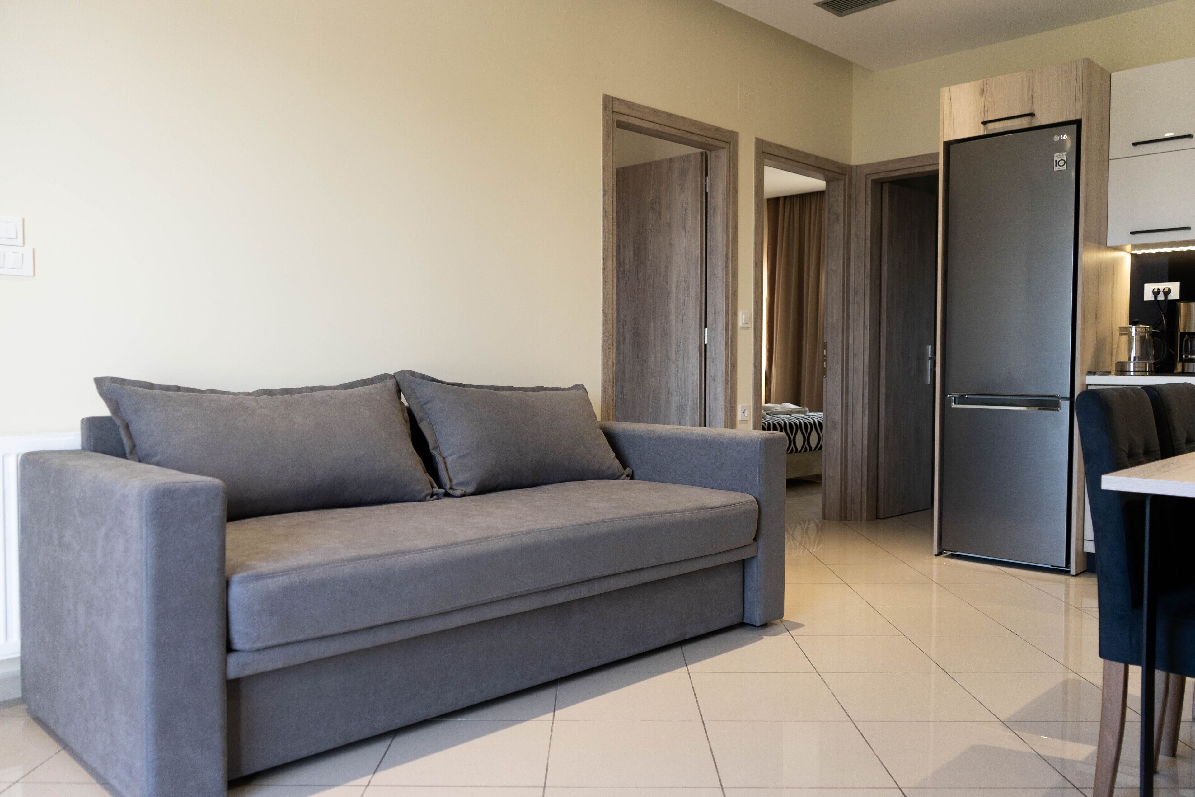 A modern living room with a grey sofa, a kitchen area with a fridge and coffee machine, and a door leading to another room.