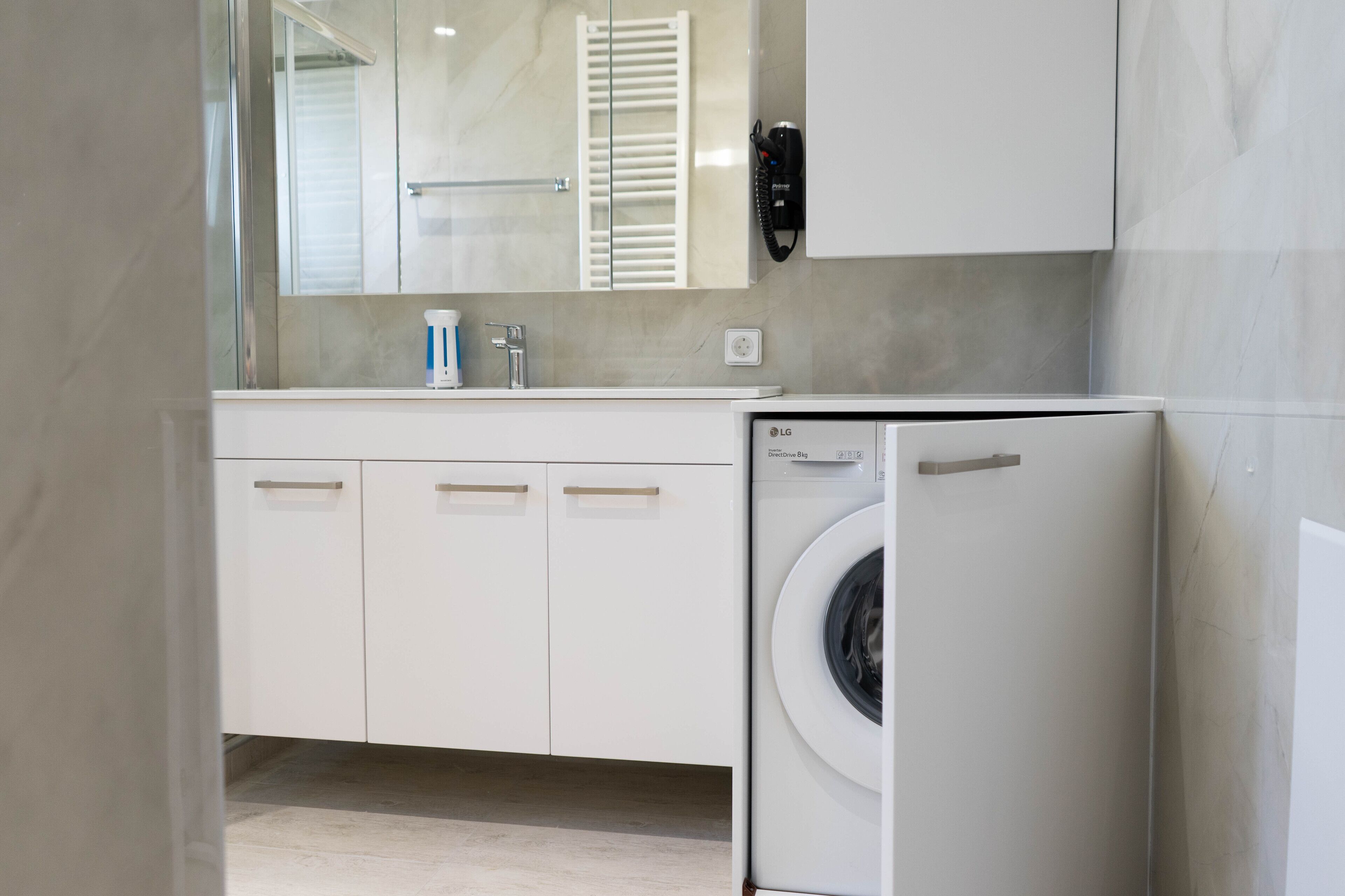 A modern bathroom with a white sink, mirror, and cabinets. There is a washing machine to the right.