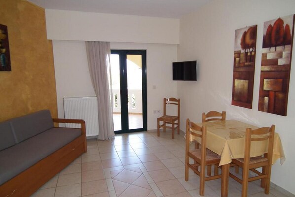 Una sala de estar con sofá, una mesa de comedor con sillas, un televisor montado en la pared y un ventanal corredero que da a un espacio exterior.