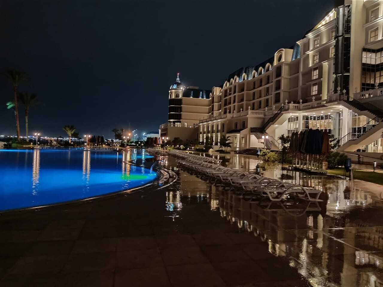 A nighttime view of a hotel with a swimming pool, reflecting pool, and a well-lit building facade.