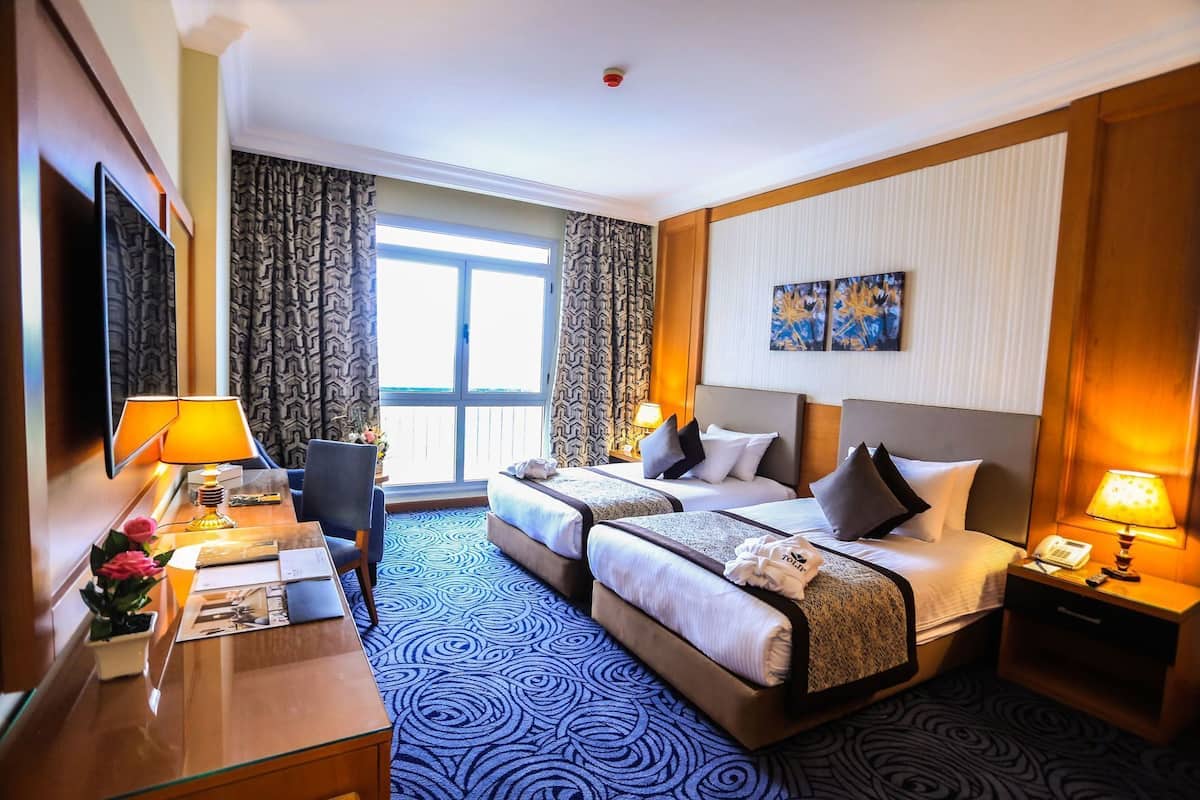 A hotel room with two beds, a desk with a lamp, a chair, a TV, and a large window.