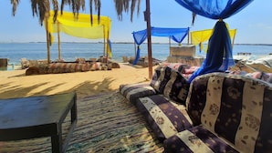 Escena de playa con sofá y sillones bajo una toldilla, una mesa de madera y vistas al mar.